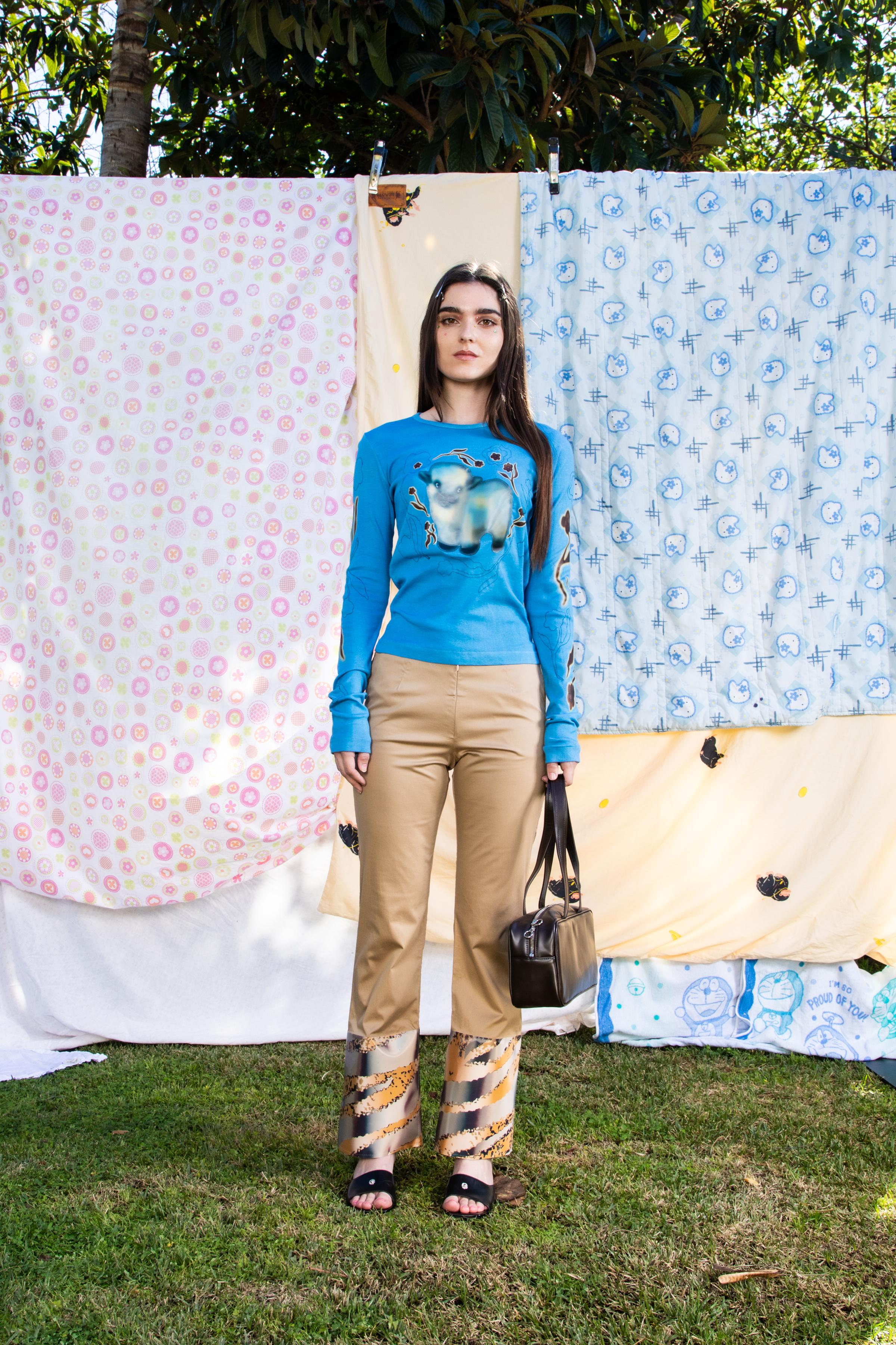 Full body image of a female model wearing a blue graphic long sleeve shirt and beige pants with a rectangular bag against a backdrop of patterned quilts.